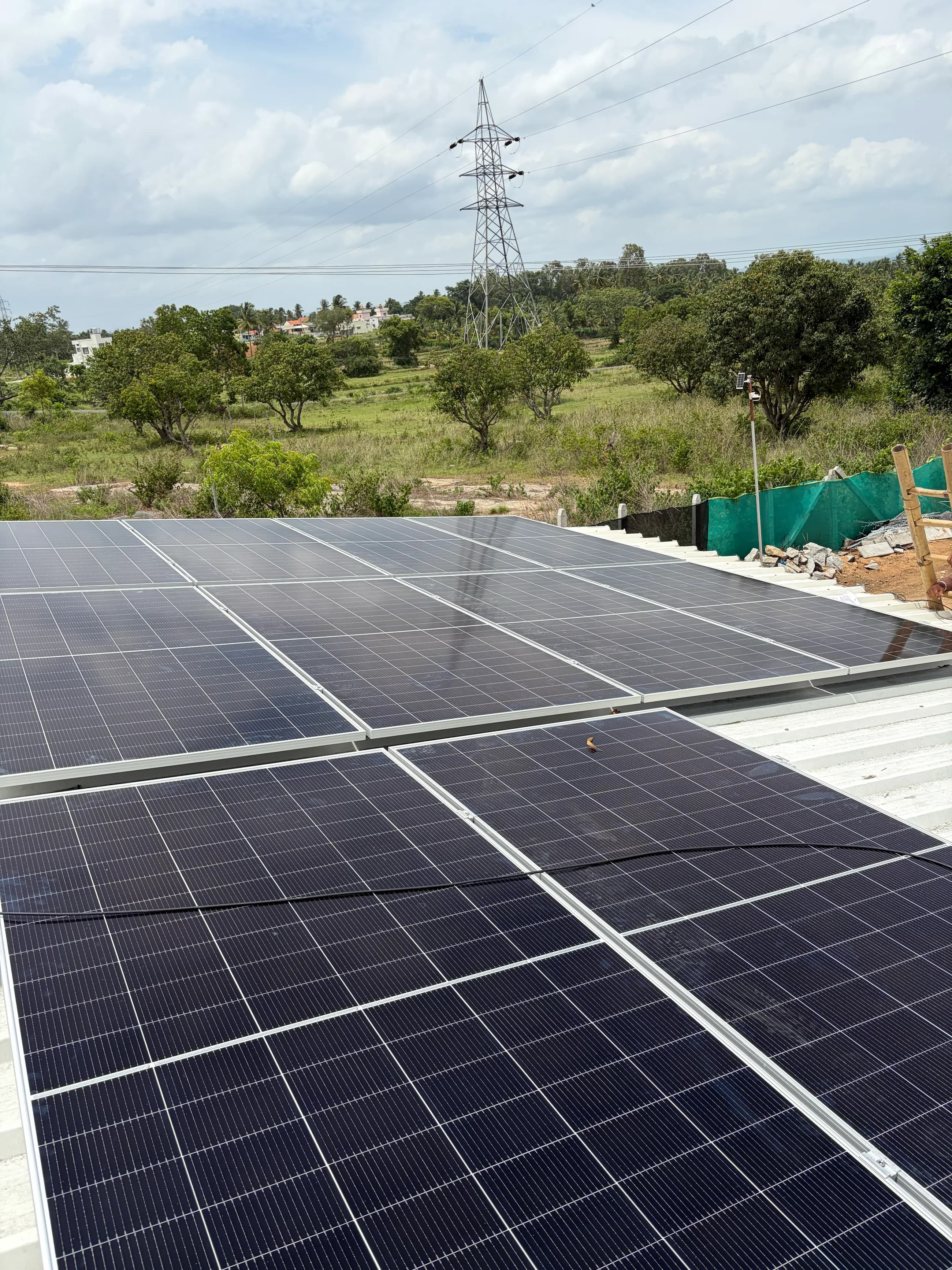 A solar array laid across a metal industrial roof, with a transmission tower, tree line and open land beyond.